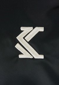 Hímzett logó fehér cérnával egy texturált fekete szövet háttérre, geometriai formákkal és letisztult vonalakkal.