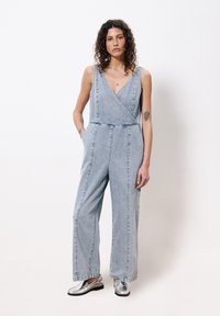 Lichtblauwe denim jumpsuit met een diepe V-hals, wijde pijpen en voorzakken. Model draagt zilveren instappers. Minimalistische stijl.