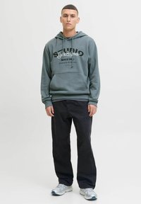 Hoodie vert foncé avec un graphisme noir à l'avant, doté de cordons de serrage et de poignets côtelés. Associé à un pantalon noir et des baskets argentées.