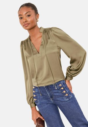 Blouse en satin vert olive avec col en V, manches bouffantes et accents à cordon, associée à un jean bleu taille haute avec des boutons dorés.