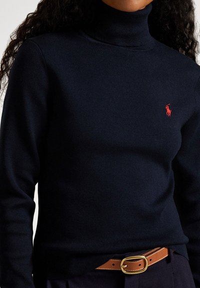 Ženska nosi temno modri pulover z visokim ovratnikom z rdečo Polo Ralph Lauren značko in rjav pas iz usnja z zlatim zaponkom.