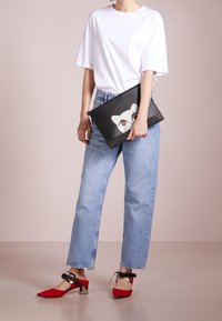 Musta clutch-laukku, jossa kissakuosi punaisilla silmillä ja kimaltavilla yksityiskohdilla, jonka henkilö valkoisessa t-paidassa ja vaaleansinisissä farkuissa pitää kädessään. Punaiset kengät.