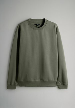 Zaļa džemperis ar apaļu kakla izgriezumu, garām piedurknēm, ribotām aprocēm un apakšmalu. Mīksta tekstūra, vienkāršs dizains bez rakstiem vai akcentiem.