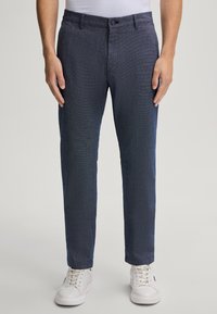 Pantaloni blu scuro con un motivo testurizzato, dotati di chiusura a bottone e tasche laterali. Abbinati a sneaker bianche per un contrasto.