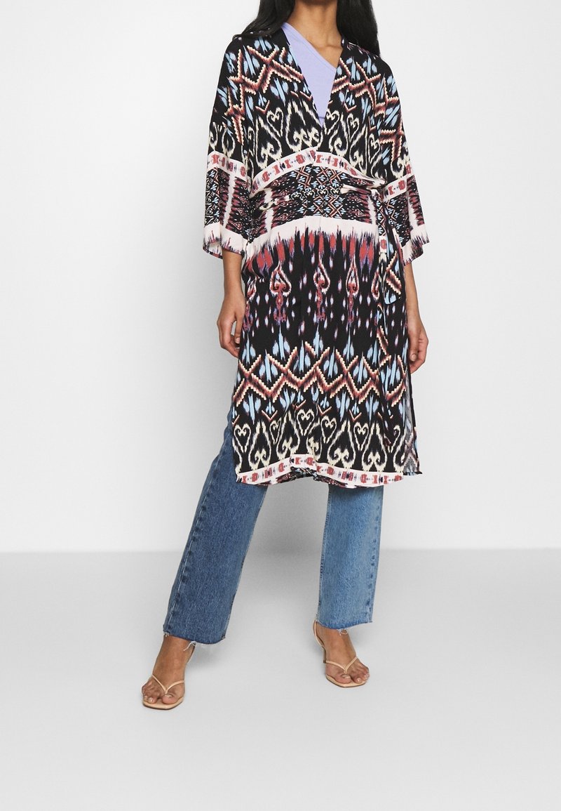 Keftan à motifs multicolores avec col en V, manches larges et ourlet à franges, accompagné d'un jean bleu à jambes larges et de sandales à brides.