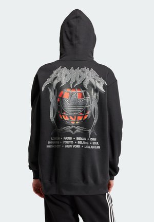 Personne portant un sweat à capuche noir avec un globe enflammé et un texte stylisé dans le dos, listant les principales villes mondiales en police argentée.