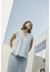 Blouse bleu clair à manches courtes avec un col froncé, portée avec une jupe en denim. L'arrière-plan présente une architecture moderne et un ciel dégagé.
