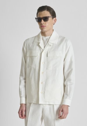 Man draagt een wit hemd met knopen en twee borstzakken en bijpassende witte broek, aangevuld met donkere zonnebril.