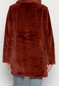 Person med en lang, blød, brun faux fur frakke set bagfra, parret med lyseblå jeans mod en ensfarvet baggrund.