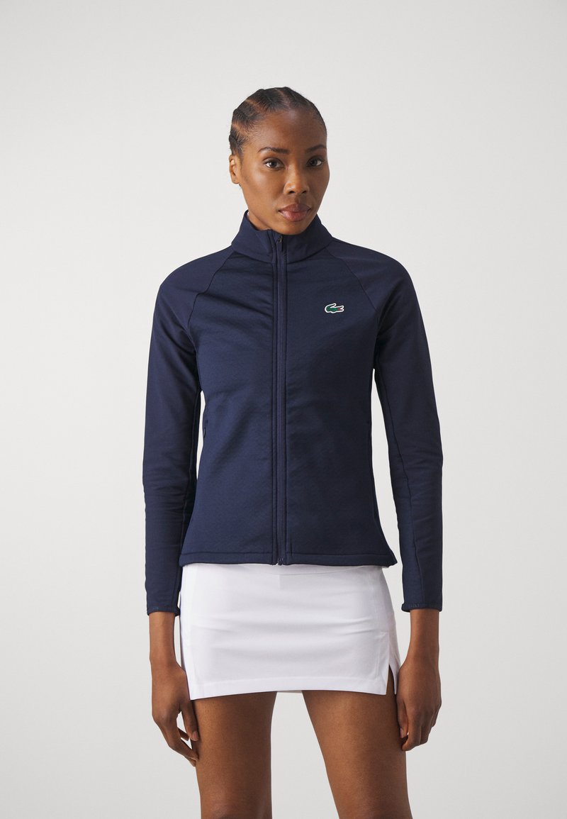 Veste zippée marine en tissu extensible, avec un col haut, des manches longues et un petit logo vert brodé sur la poitrine.