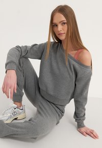 Hall tõmblusega hall sweatshirti off-shoulder disain, koos sobivate joggeritega. Mudel kannab valgeid sportlikke tossusid hallide aktsentidega.