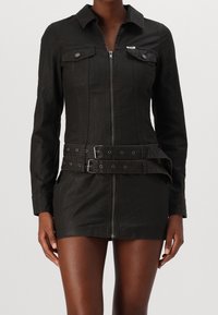 Svart denimklänning med långa ärmar, dragkedja fram, bröstfickor och dubbel spännbak vid midjan. Slät textur och figurnära passform.