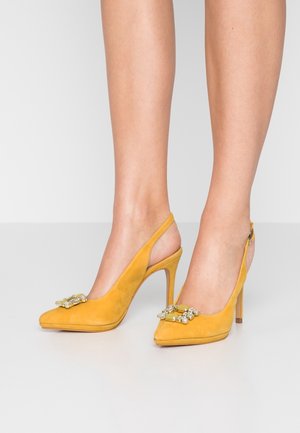 Żółte zamszowe szpilki typu slingback z szpiczastymi noskami i ozdobnymi klamrami z biżuterią na stopach na jednolitym tle.