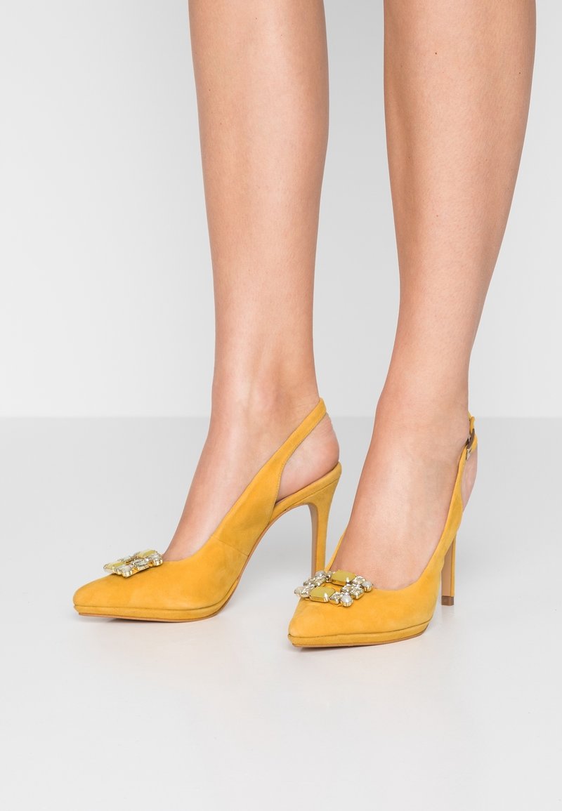 Escarpins slingback en daim jaune à bouts pointus avec boucles ornées de bijoux portés sur des pieds, sur un fond uni.