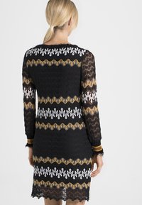 Svart klänning med långa ärmar som har strukturerade mönster i gult, vitt och guld. Nederkant och ärmslut har kontrasterande ränder.