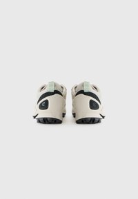 Helle beige Sportschuhe mit schwarzen Akzenten und strukturierten Seiten. Verfügt über eine grüne Schlaufe und eine Gummisohle mit ausgeprägtem Profil.