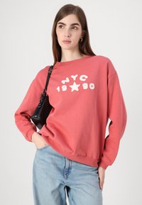 Sudadera coral con estampado blanco de NYC 1990, cuello redondo, hombros caídos y corte relajado. Combinada con jeans de color azul claro y un bolso negro pequeño.