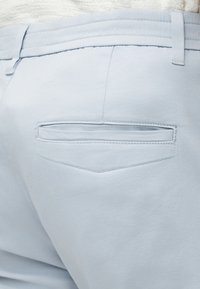 Nærbillede af lyseblå bukser med baglomme med vandret slids og bæltestropper, båret med en hvid tekstureret top.