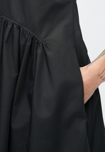 Fekete ruha, melynél a derékrészen összegyűrt anyag voluminózus sziluettet alkot. Az anyag simának, matt hatásúnak tűnik.