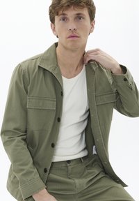 Olivgrüne, langärmelige Jacke mit Knopfschluss, zwei Brusttaschen, getragen über einem weißen, gerippten Tanktop, kombiniert mit passenden Hosen.
