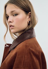 Veste en daim marron avec un col en cuir marron foncé. La veste a un design épuré et une texture subtile, avec un détail de crochet en argent.
