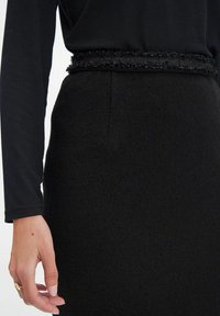 Jupe noire avec un tissu texturé et une taille effilochée, associée à un haut noir à manches longues. Main reposant doucement sur la jupe.