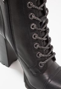 Botín de cuero negro con un alto tacón de bloque, que presenta ojetes plateados y cordones negros y plateados, con costuras detalladas a lo largo de los lados.