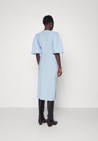 Abito azzurro chiaro con maniche a sbuffo, taglio al ginocchio e silhouette aderente. Indossato con stivali neri fino al ginocchio. Tessuto liscio.
