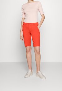 Kvinna i en ljusrosa kortärmad topp, klara orange knälånga shorts och vita sneakers, stående mot en enkel bakgrund.