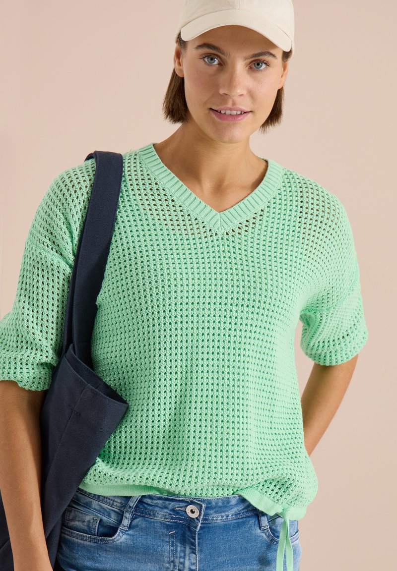 Femme portant un pull en maille vert menthe, un jean bleu, une casquette beige et portant un sac cabas bleu marine sur l'épaule devant un fond beige.