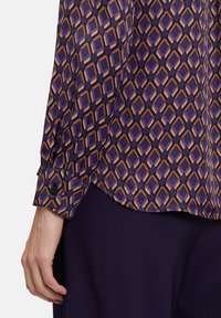 Patterned blouse featuring a purple, black, and orange geometric design. Long sleeves with buttoned cuffs, silky texture, and a curved hem.