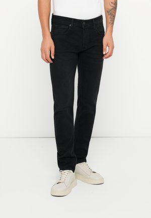 Homme portant un jean noir slim, des baskets blanches et une chemise blanche, debout devant un fond blanc uni.