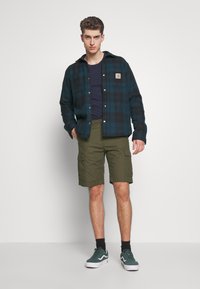 Camisa de franela a cuadros en verde oscuro y negro, superpuesta sobre una camiseta de color marino. Pantalones cortos cargo en verde oliva, calcetines oscuros y zapatillas verdes completan el conjunto.