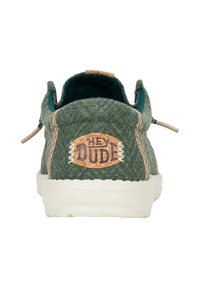 Scarpa in tessuto verde con superficie texturizzata, suola bianca e un'etichetta circolare in sughero con la scritta "HEY DUDE", caratterizzata da dettagli e lacci beige.