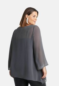 Blusa grigia trasparente con maniche a campana lunghe, orlo decorato con piccole scintille, texture morbida e taglio asimmetrico.