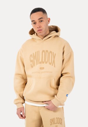 Ung mand iført beige SMILODOX hoodie og matchende joggingbukser, stående mod en ensfarvet lys baggrund.