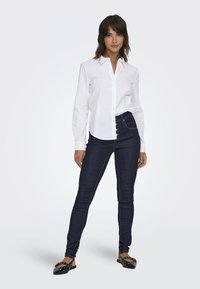 Witte blouse, donkerblauwe skinny jeans en zwarte ballerina's met studs. De blouse heeft lange mouwen en een klassieke kraag.