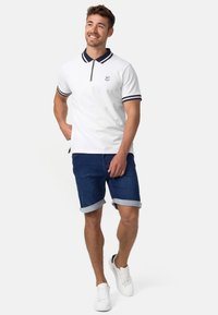 Bijela polo majica s ovratnikom na zip i tamnoplavim detaljima, uparena s poderanim traper šortsevima. Model nosi bijele tenisice, što stvara ležeran izgled.