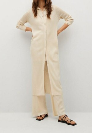 Frau trägt eine cremefarbene, gerippte lange geknöpfte Strickjacke und passende weite Hosen, kombiniert mit schwarzen flachen Sandalen mit Riemchen.