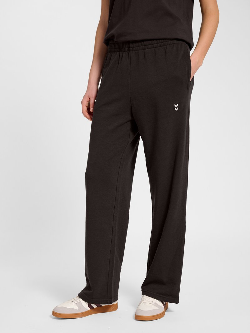 Personne portant un pantalon de survêtement noir ample avec un logo chevron blanc sur la cuisse gauche et des baskets blanches avec des semelles marron.