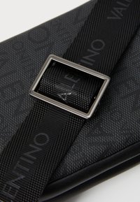 Sac noir texturé avec motif de logo, doté d'une bandoulière à tissage et d'une boucle carrée en argent. Le matériau est durable et élégant.