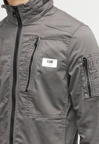 Szara kurtka na suwak z wieloma kieszeniami, z kołnierzem typu stójka i naszywką z logo. Wykonana z gładkiej, trwałej tkaniny o teksturowanym wykończeniu.