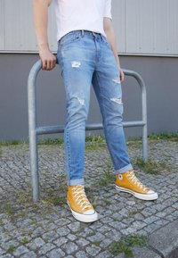 Persona con vaqueros rasgados y zapatillas altas amarillas apoyada en un aparcamiento de bicicletas de metal sobre un pavimento de adoquines.