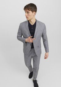 Completo grigio chiaro su misura con finitura testurizzata, chiusura a un bottone e pantaloni coordinati. Indossato sopra una maglietta polo scura. Scarpe nere.