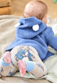 Zils adīts kapuci džemperis ar baltām ausu akcentiem, salikts ar bēšām legingu, uz kuriem attēlots zils zaķis, uz neitrāla segas.