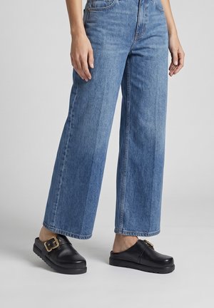 Persoon die wijde blauwe jeans en zwarte leren instapklompen met gouden gespen draagt, staand op een lichtgrijze vloer.