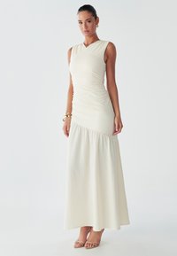 Le mannequin se tient avec assurance dans une robe crème ajustée sans manches, avec une jupe évasée, mettant en valeur de belles sandales et accessoirisée avec des bijoux en or.