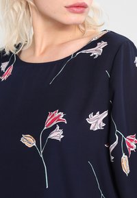 Blusa blu navy con scollatura tonda, caratterizzata da motivi floreali in rosso, rosa e giallo su un tessuto liscio. Maniche lunghe con fantasie floreali.