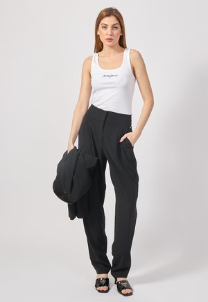 Top bianco senza maniche con testo in nero, pantaloni neri a vita alta e ciabatte nere con dettagli del logo, mentre tiene una giacca nera.
