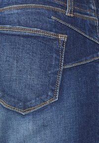 Jeans de ganga de cor azul escura com um bolso traseiro com costura amarela em contraste e uma aparência de tecido texturizado.
